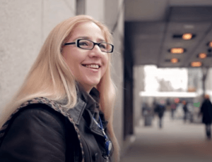 Diana sits on a bench looking just past the camera to her right. She has long straight blonde hair and wears black-rimmed glasses.