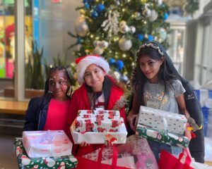 Three young girls standing in front of a Christmas tree, holding holiday gifts.