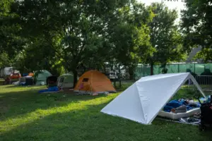 A lawn with half a dozen tents that sit below a handful of trees. Under the first tent, closest to the camera, two people rest on a foam mattress.