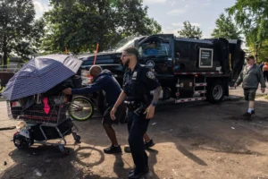 Un miembro del departamento de policía de Nueva York camina junto a un individuo que empuja un carrito de compras con varias pertenencias personales, cubierto por un paraguas. Al fondo hay un camión de Parques de la Ciudad de Nueva York y está situado en medio de un claro.