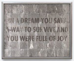 A conceptual artwork that features a textured silver surface with a reflective quality, on which the text 'IN A DREAM YOU SAW A WAY TO SURVIVE AND YOU WERE FULL OF JOY' is boldly displayed in metallic capital letters.