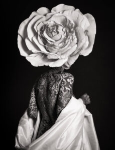 This black-and-white photograph features a figure with intricate full-body tattoos, draped in a flowing white fabric. The figure's head is replaced by an enormous, highly detailed rose blossom.