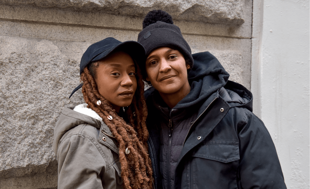 A couple stand close to each other in front of a sandstone wall. They are wearing winter coats, one has her hair in locks and dons a cap while the other wears a beanie. Their faces are somber, and expressions neutral. Image credit: Seze Devres