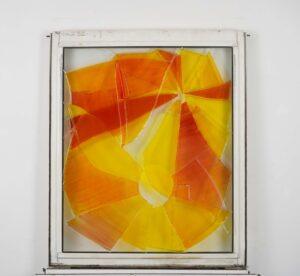 This stained glass artwork features a vibrant abstract design with large, angular shards of glass in vivid hues of orange, yellow, and red. The stained glass is set within a weathered white frame, enhancing its raw and hand-crafted aesthetic.