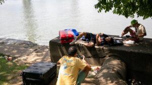 Jóvenes en pantalones cortos y camisetas descansan bajo la sombra de un árbol, junto a un río. Llevan consigo su equipaje.