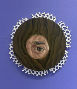 A circular painting featuring a realistic human eye peering through a small hole in a dark wood-like surface. The detailed eye is surrounded by a textured pattern resembling wood grain, with the painting bordered by a white crochet doily.