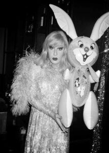 A black-and-white photograph from January 1986 featuring drag icon Lady Bunny, dressed in a gown with feathered accents and heavy makeup, holding a large inflatable bunny. The inflatable bunny has exaggerated cartoonish features with big ears and a smiling face. Lady Bunny's expression is slightly wide-eyed and dramatic, adding to the theatrical feel of the image.