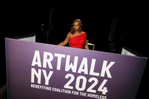 A DJ wearing a red dress in a both behind a sign that reads "ARTWLK NY 2024 benefiting Coalition for the Homeless".